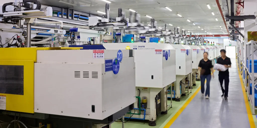 Two people walk through a row of plastic moulding machines on the production floor.